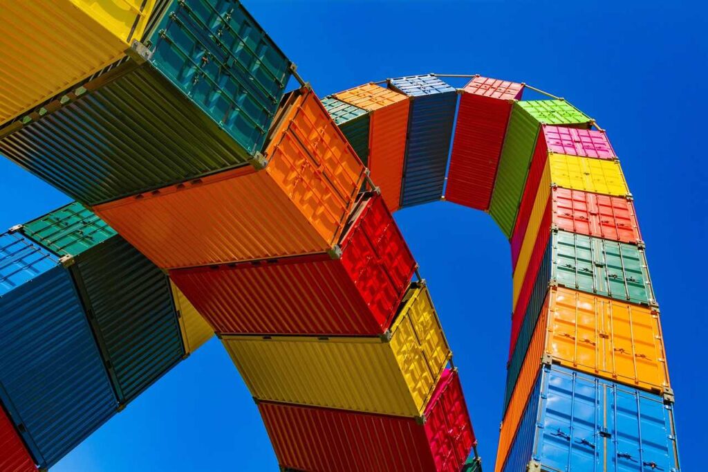 A vibrant column of shipping containers stacked neatly, showcasing a mix of bright and bold colors such as red, blue, yellow, and green, with some containers featuring logos and markings. The scene reflects a busy industrial or port setting, emphasizing organization and global trade.